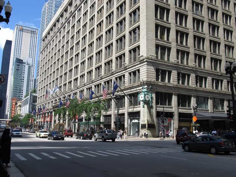 image from across the street of a large tall tan building with a green exterior clock hanging from its corner